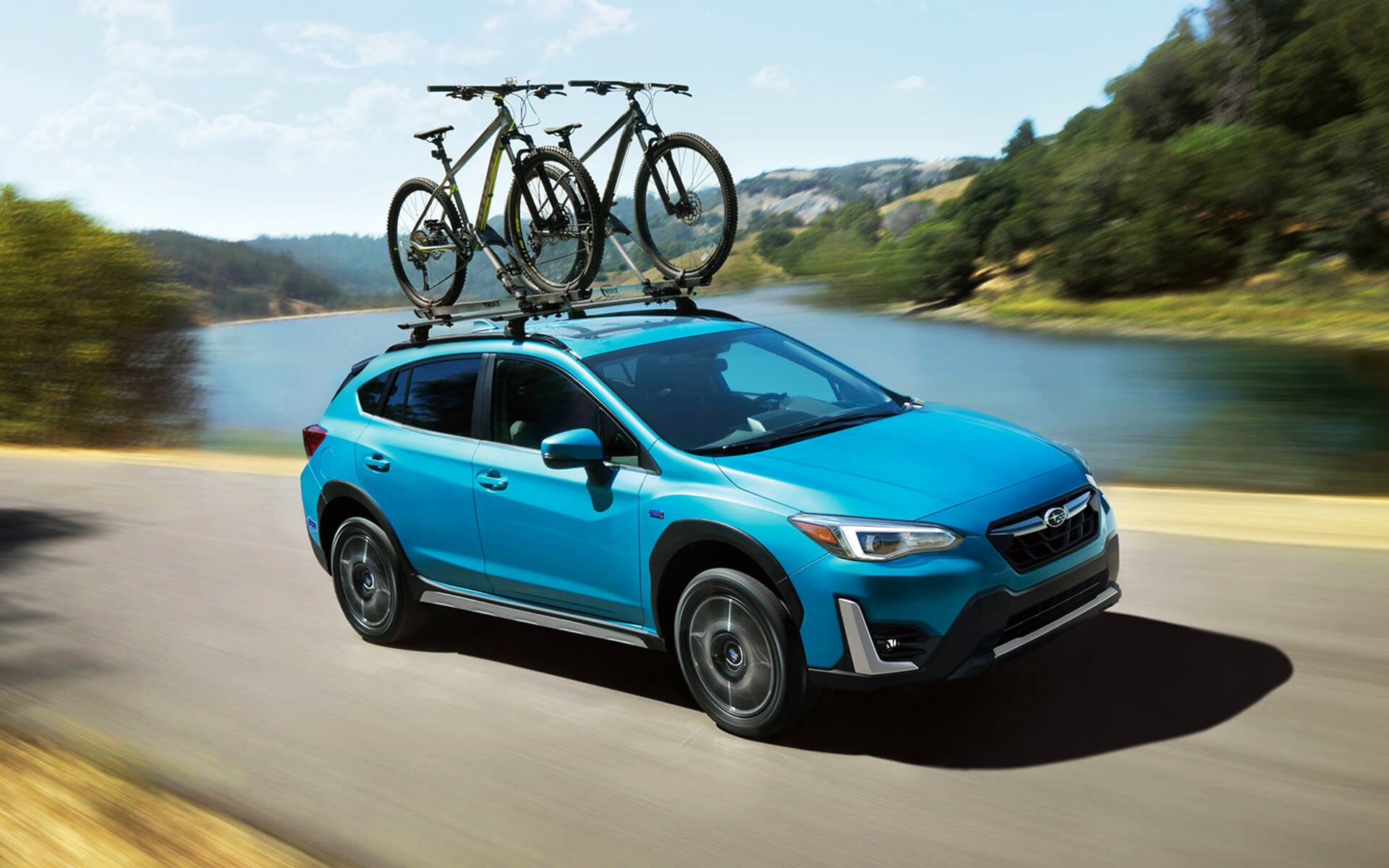 A blue Crosstrek Hybrid with two bicycles on its roof rack driving beside a river | Five Star Subaru in Grapevine TX
